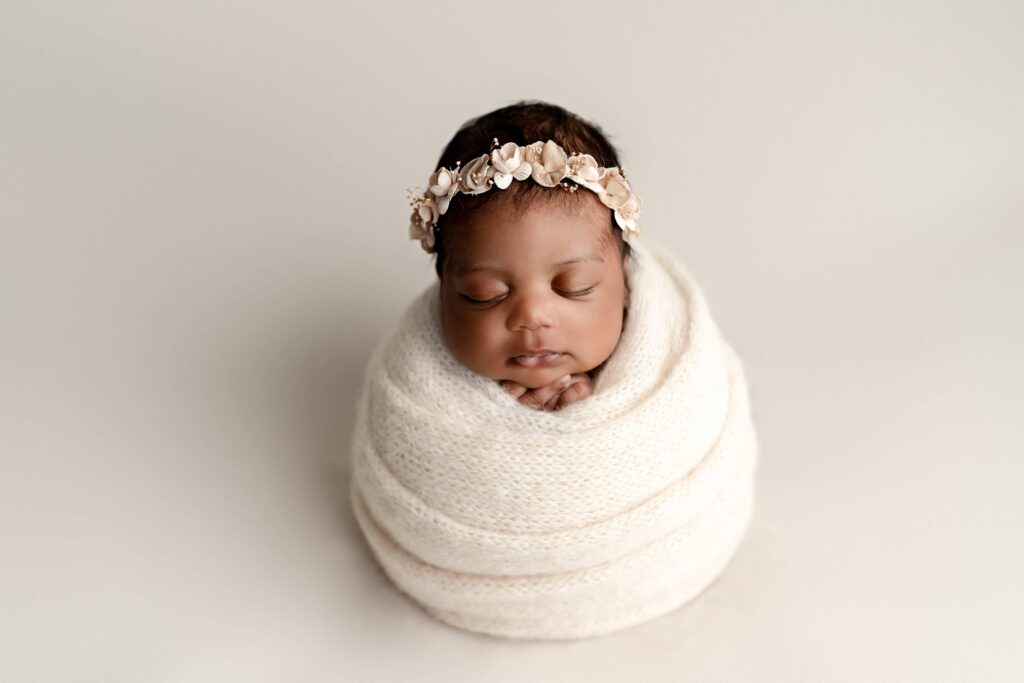 A beautiful newborn baby girl peacefully sleeping, wrapped in a cream wrap with flower headband. 