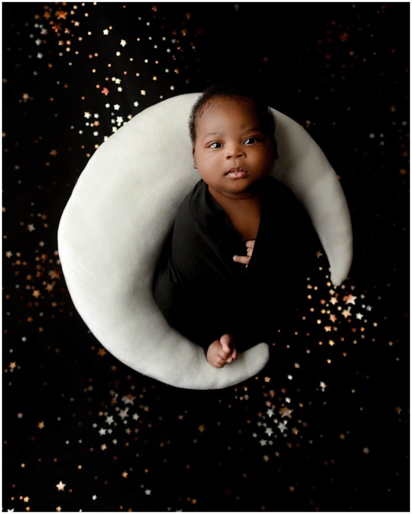 Little baby boy wrapped up in black fabric with toes and hands out. There is a backstop with stars and a moon prop pillow that baby is laying on.