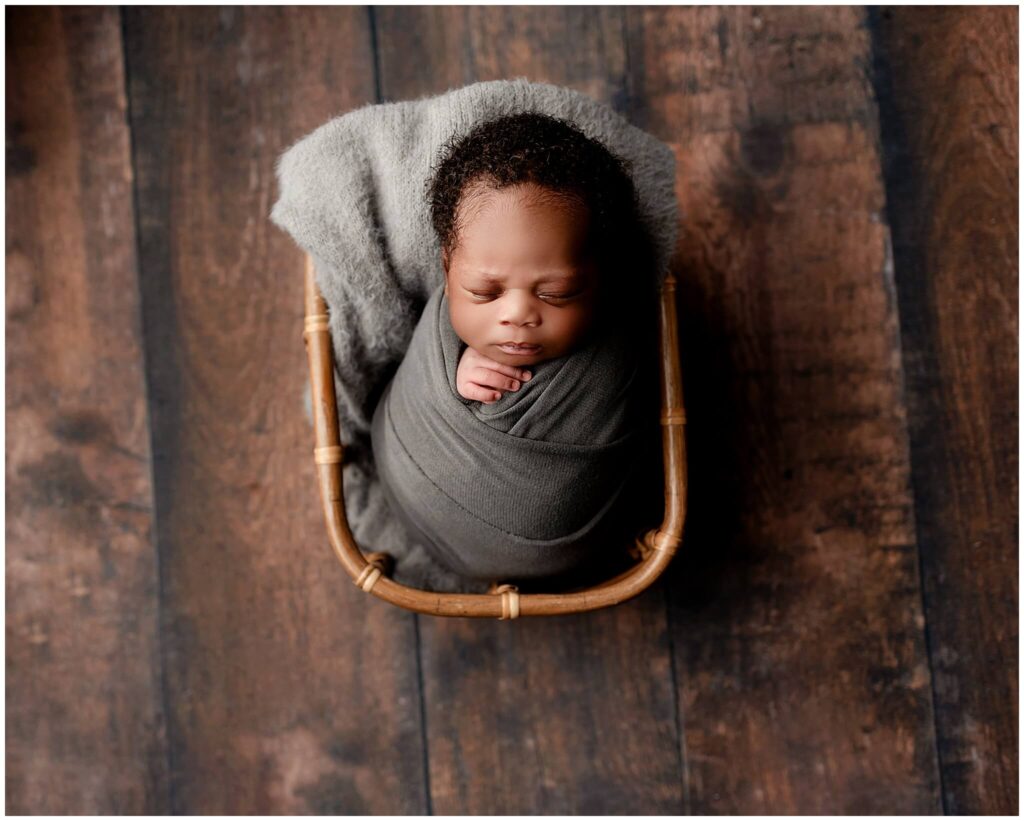 Newborn prop basket with baby boy wrapped in gray fabric and laying in the basket sleeping.