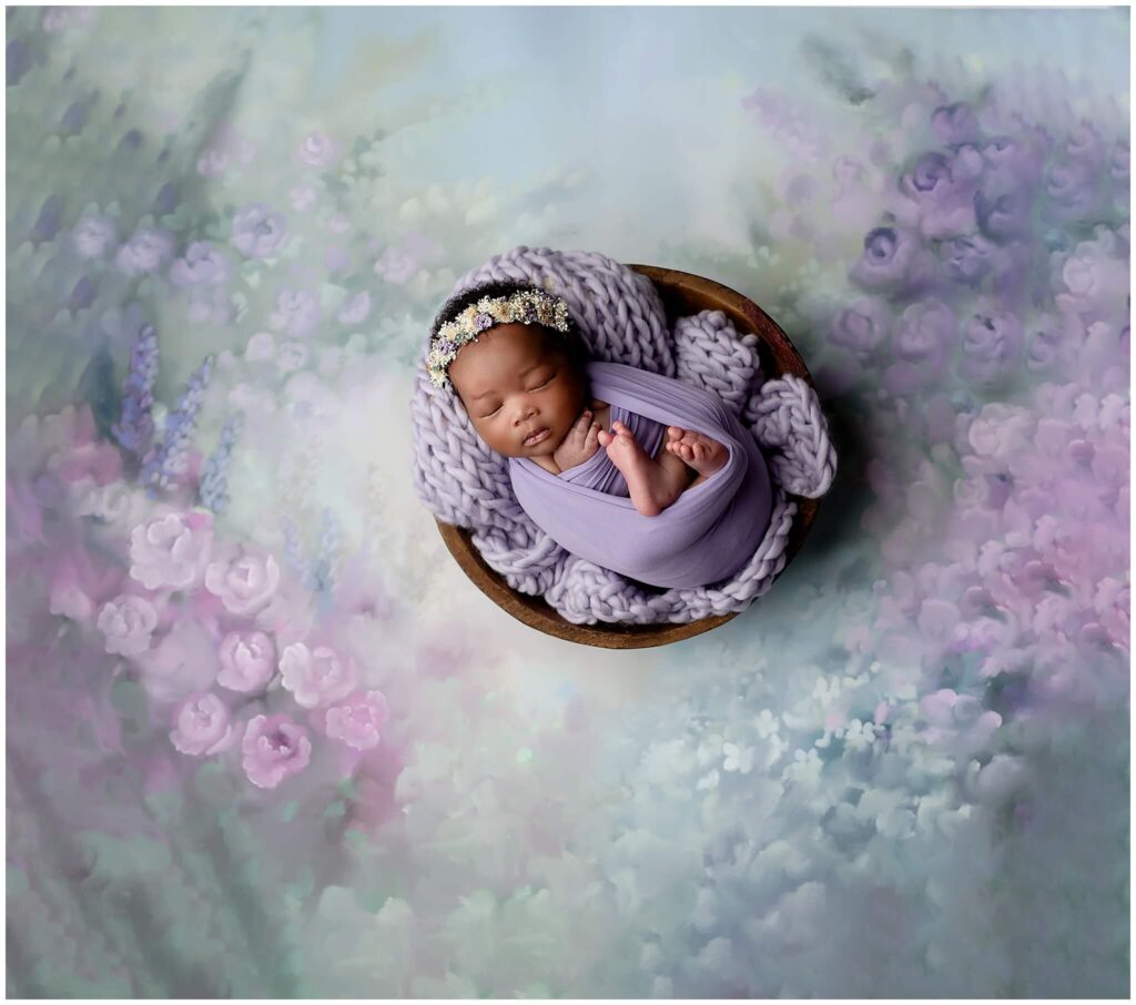 Newborn baby wrapped in purple wrap sleeping in a bowl prop. The prop is on a beautiful floral backdrop.
