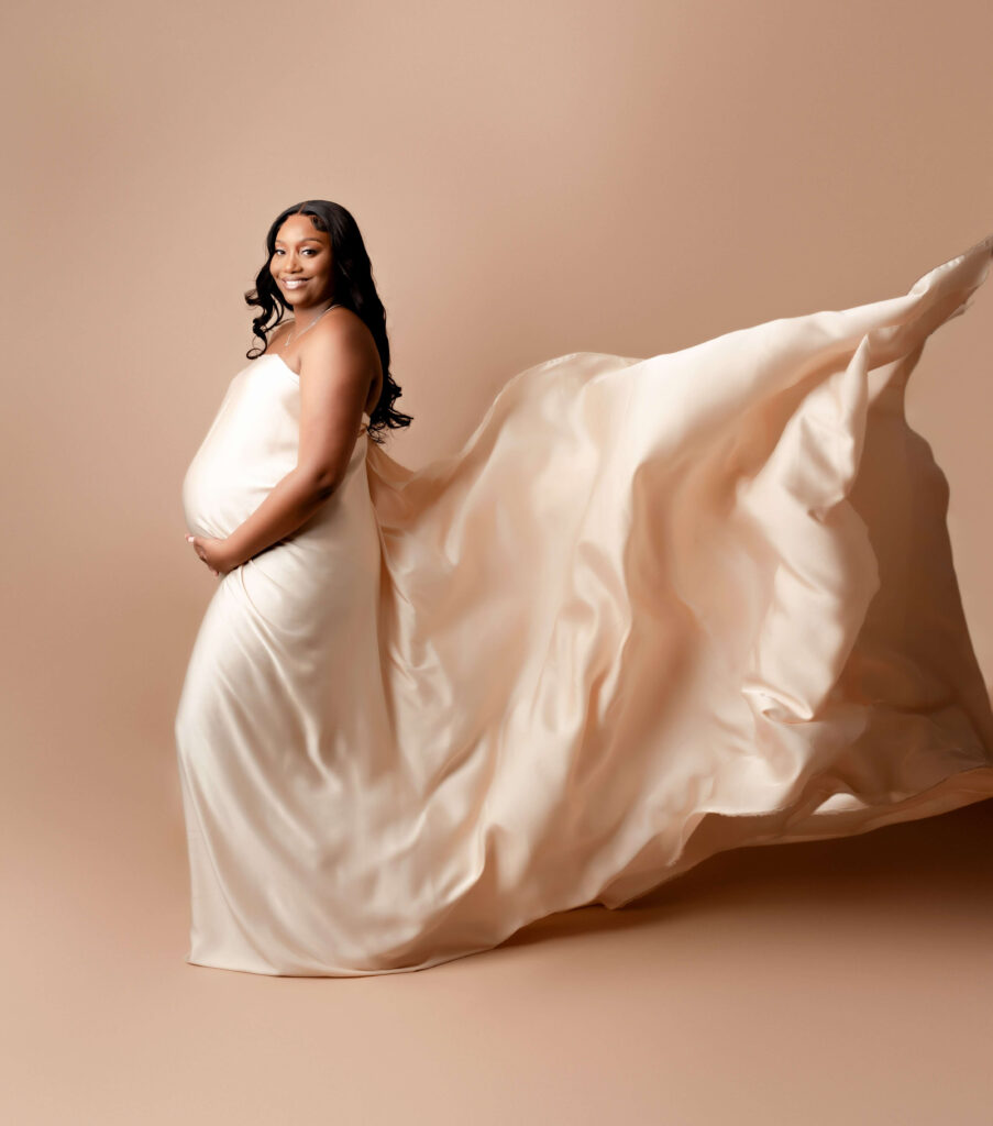 A pregnant woman standing in front of backdrop, wearing a flowing piece of fabric. It is billowing in the wind behind her, creating a beautiful and ethereal effect.