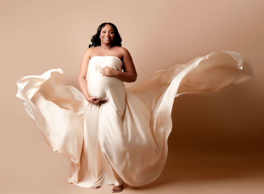 A pregnant woman standing in front of backdrop, wearing a flowing piece of fabric. It is billowing in the wind, creating a beautiful and ethereal effect.