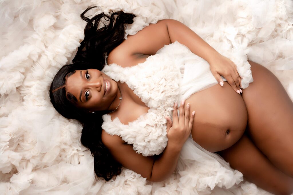 A pregnant woman lying on the floor in a bed of fluffy fabric, cradling her baby bump with a huge smile on her face.