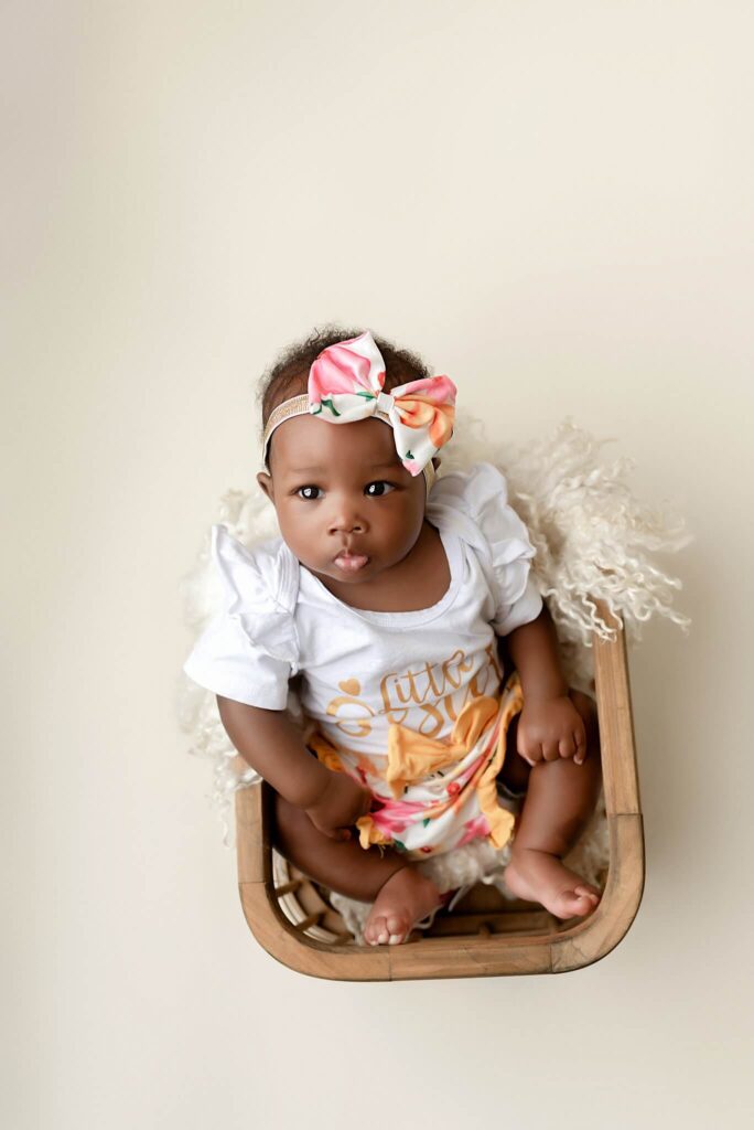3 month old baby girl wearing a cute peach floral outfit with headband. She is sitting in a basket.