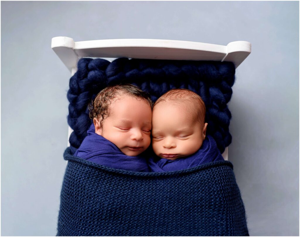 Twins laying cozy together all wrapped up under a blanket on a white little bed.