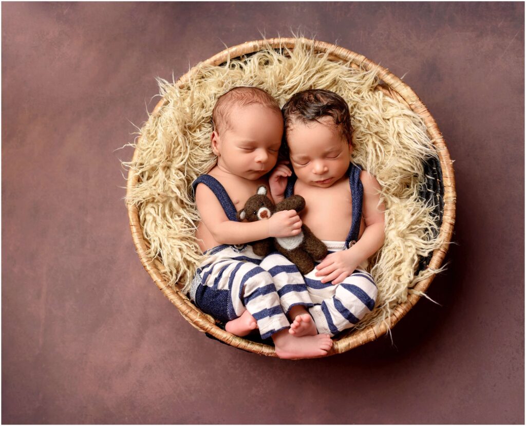 A pair of newborn twins holding a teddy bear while sleeping all curled up in a basket.