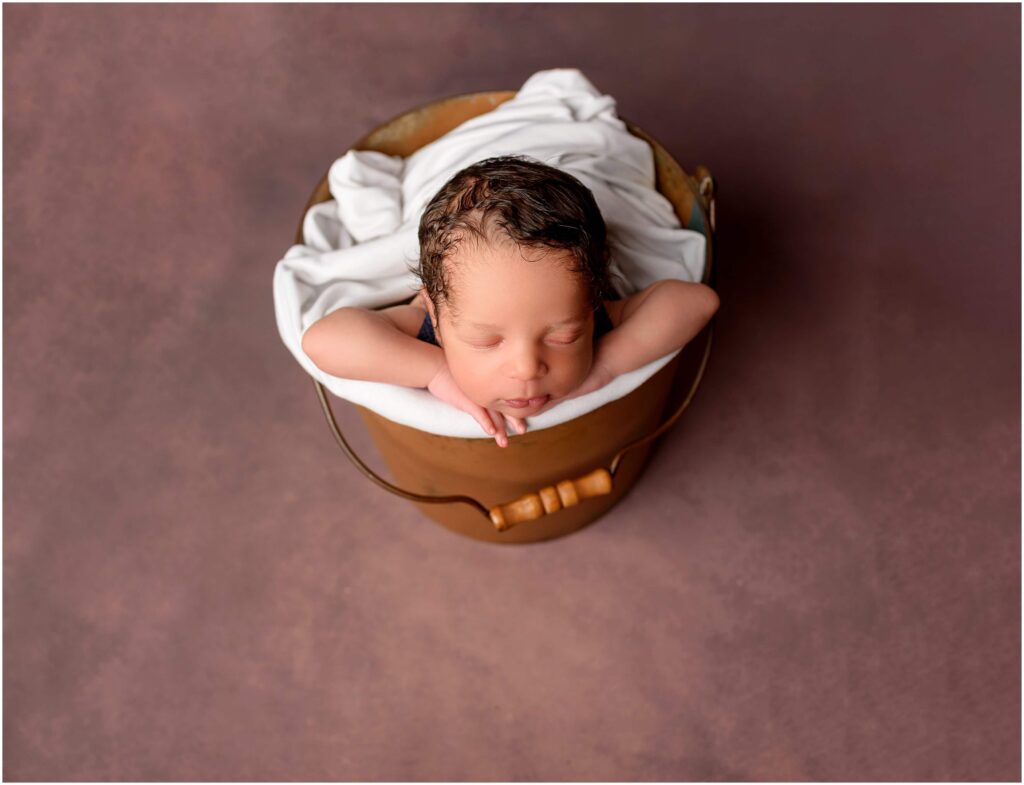 Newborn boy posed in a bucket with chin on hands sleeping wrapped in white fabric.