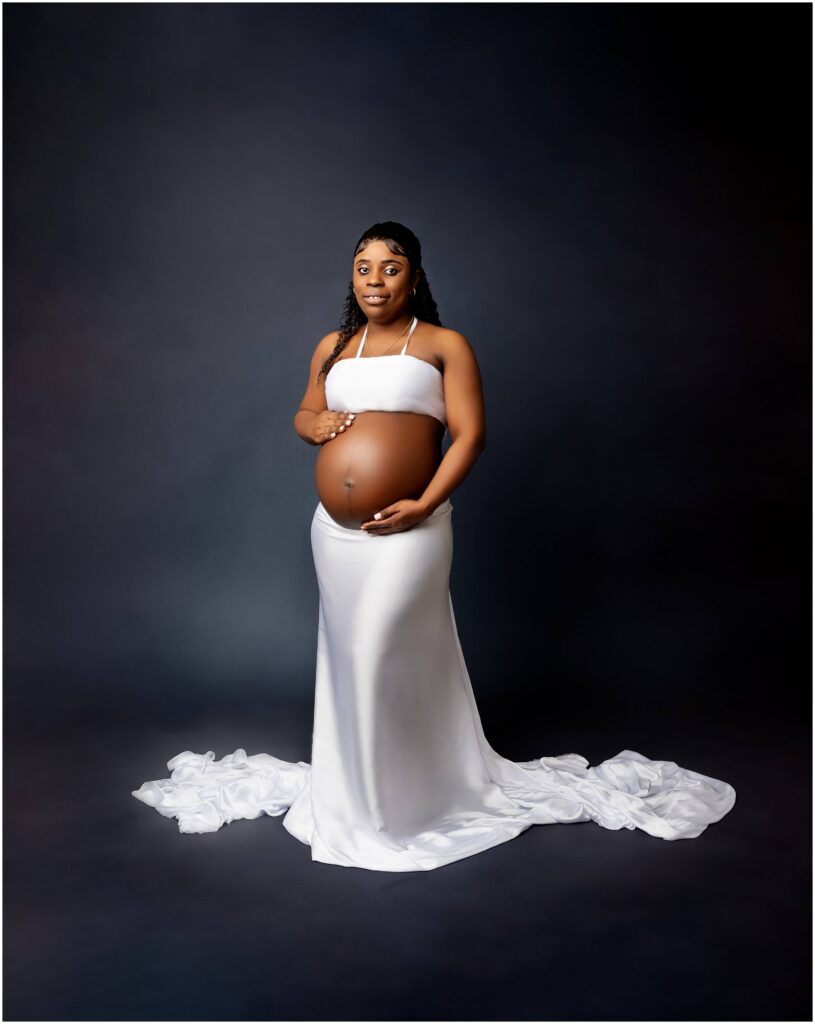 White wrap outfit on pregnant woman holding baby bump, standing on blue backdrop. 