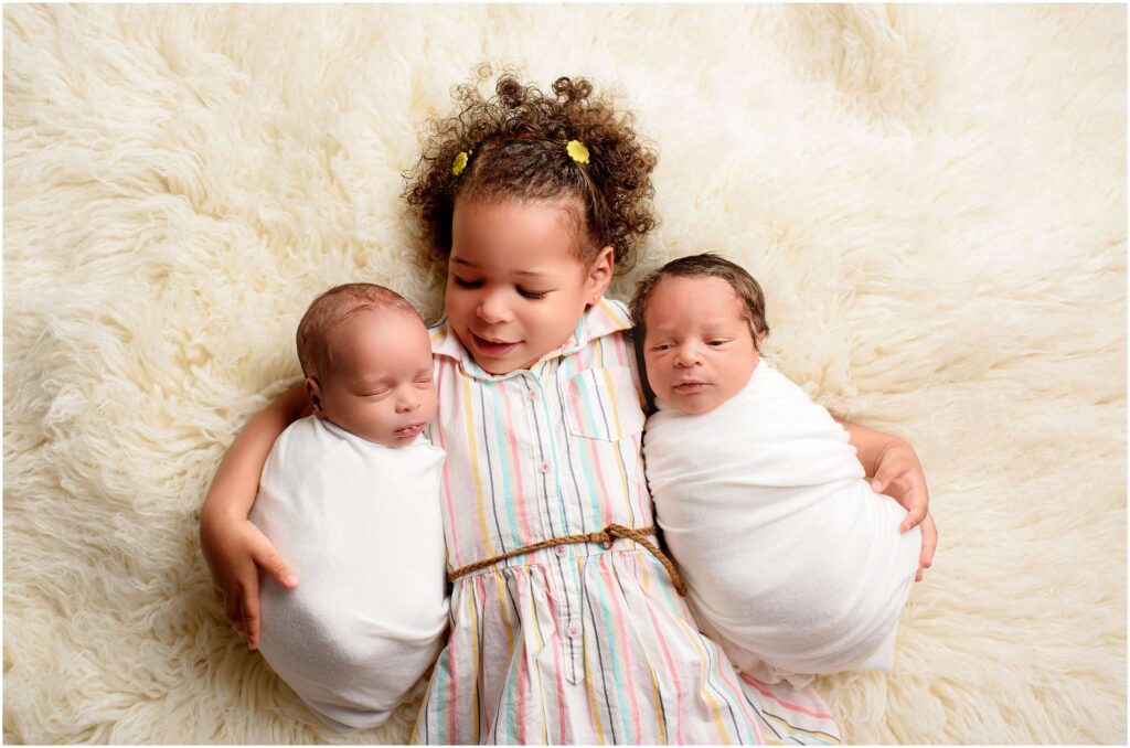 Newborn twins snuggled next to their sister while she is looking at one of them sleeping and the other one is awake.